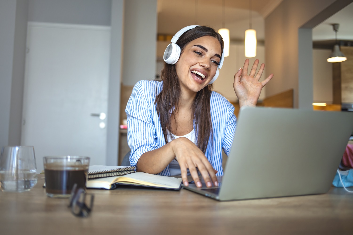 Smiling girl student wear wireless headphone study online with skype teacher, happy young woman learn language listen lecture watch webinar write notes look at laptop sit in cafe, distant education